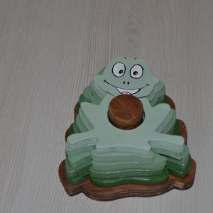 Peut inclure: Un jouet d'empilage en bois en forme de grenouille avec une finition peinte en vert et un visage souriant. La grenouille est composée de plusieurs couches de bois, chacune peinte dans une nuance de vert légèrement différente.