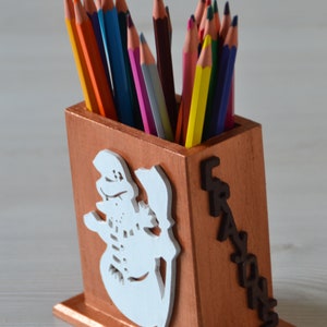 Può includere: Un porta matite in legno con un disegno di pupazzo di neve ritagliato in bianco. Il porta matite è dipinto di un colore marrone chiaro e ha la parola "Crayons" scritta sul lato in lettere marroni scure. Il porta matite è pieno di matite colorate.