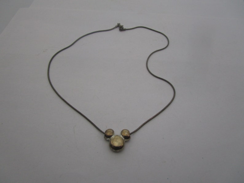 以下が含まれることがあります： 銀色のチェーンネックレスに、金色のミッキーマウスの頭のペンダントが付いている。