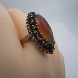 May include: A vintage ring with an oval, amber-coloured stone. The stone is framed by a dark, spiky metal setting. The ring's band is gold-toned. The ring is displayed against a light background.