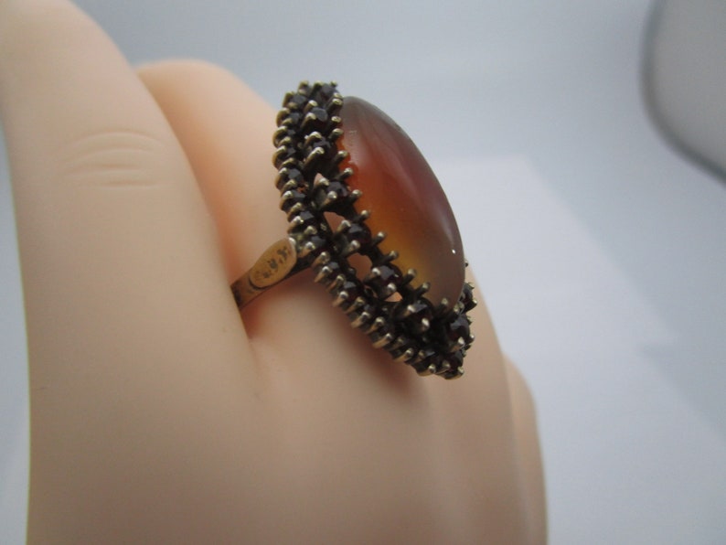 May include: A vintage ring with an oval, amber-coloured stone. The stone is framed by a dark, spiky metal setting. The ring's band is gold-toned. The ring is displayed against a light background.