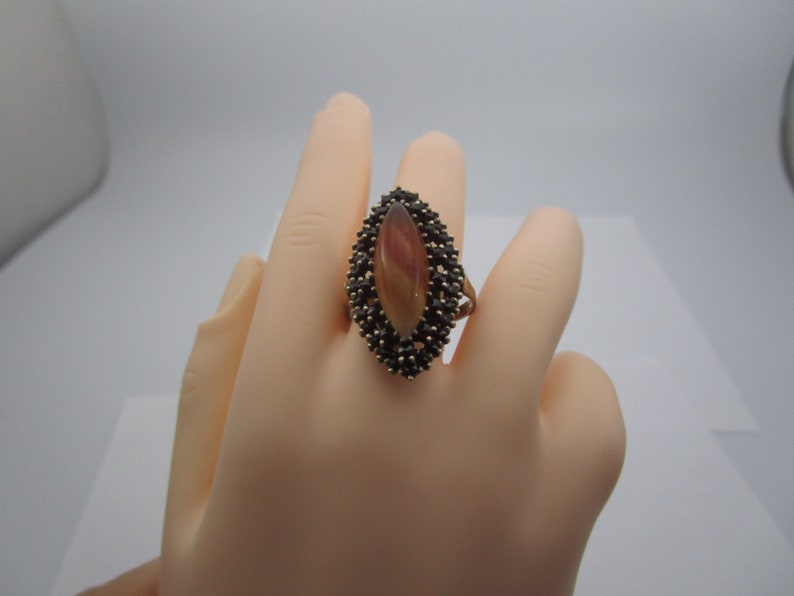 May include: A gold ring with an oval, reddish-brown and cream-coloured stone. The stone is encircled by small, dark stones. The ring is shown on a mannequin hand. The ring's design is eye-catching.