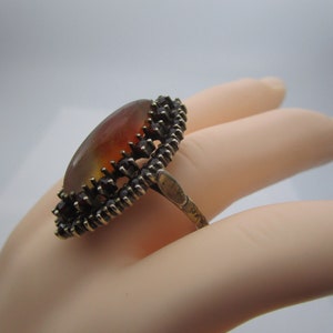 May include: An oval ring with an amber-toned stone, displaying a gradient effect. The stone is set within a dark metal frame, accented with small, dark red details. The ring's band is crafted from the same dark metal.