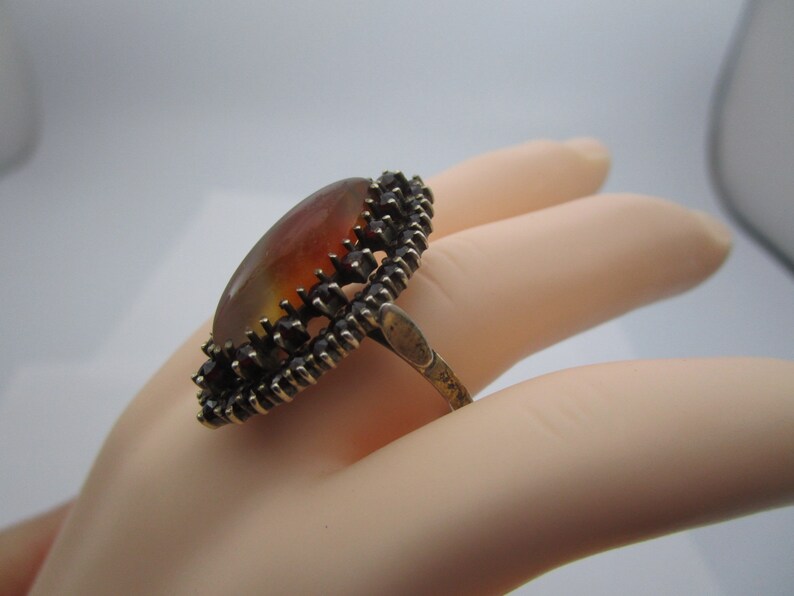 May include: An oval ring with an amber-toned stone, displaying a gradient effect. The stone is set within a dark metal frame, accented with small, dark red details. The ring's band is crafted from the same dark metal.