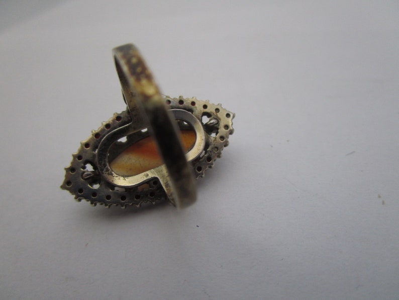 May include: A vintage silver ring featuring an oval, light orange stone. The stone is encircled by small, clear stones. The ring's band has a simple, thin design.