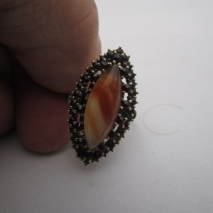 May include: An oval ring with a central, elongated orange and white stone, encircled by small, dark gemstones. The ring has a gold-coloured setting, providing a contrast with the darker stones. A piece of jewellery.