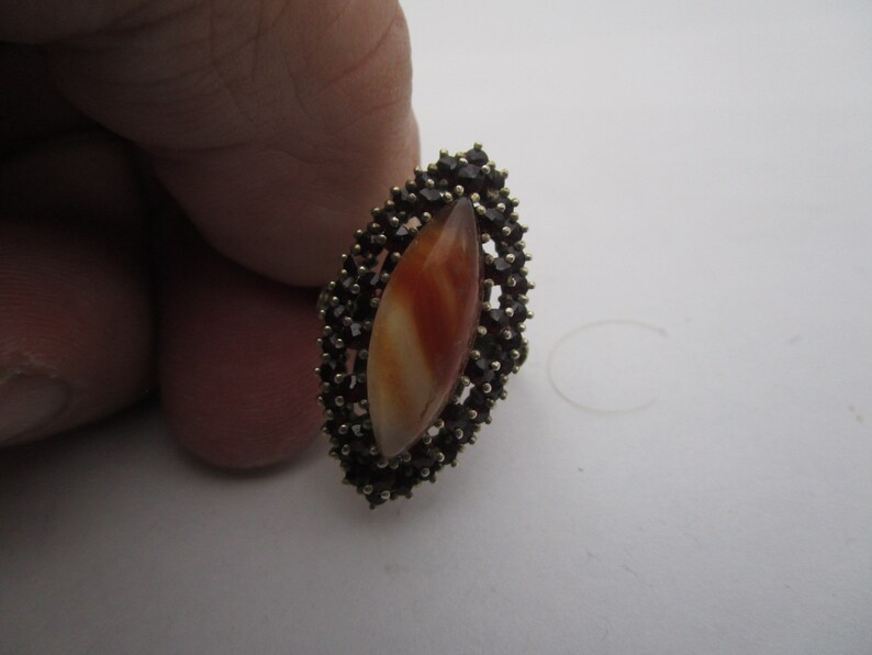 May include: An oval ring with a central, elongated orange and white stone, encircled by small, dark gemstones. The ring has a gold-coloured setting, providing a contrast with the darker stones. A piece of jewellery.