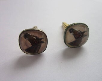 May include: A pair of silver cufflinks with a brown horse head design. The horse has a red eye and is set against a white background.