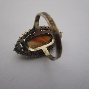 May include: A vintage silver ring with an oval, orange-brown stone. The stone is encircled by small, red-orange gemstones and a silver setting. The ring's band is also silver.