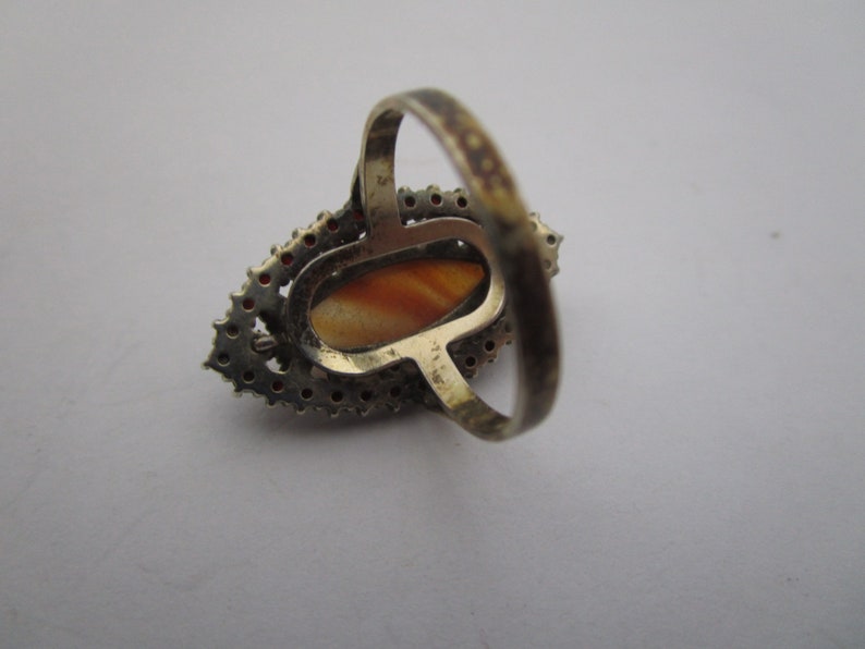 May include: A vintage silver ring with an oval, orange-brown stone. The stone is encircled by small, red-orange gemstones and a silver setting. The ring's band is also silver.
