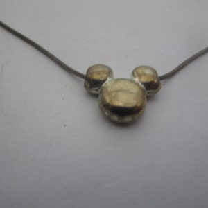 以下が含まれることがあります： 銀色のチェーンネックレスに、金色のミッキーマウスの頭のチャームが付いている。