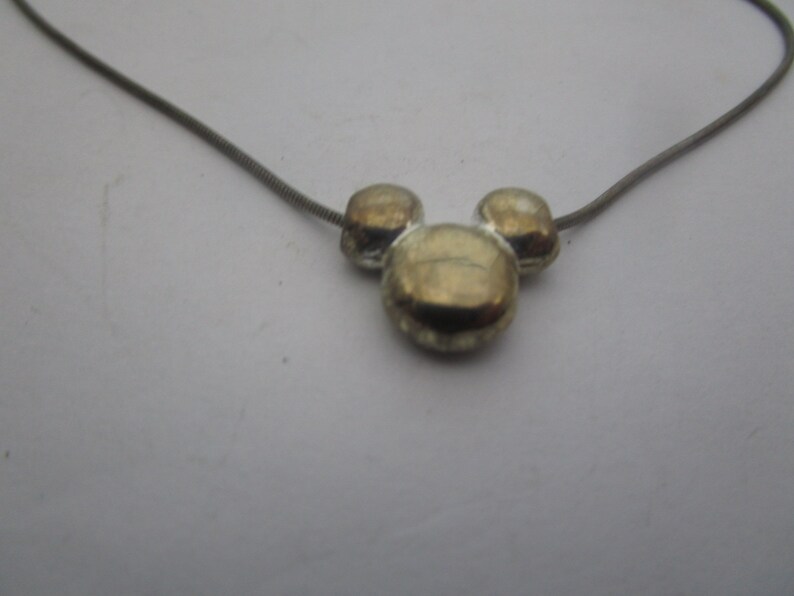 以下が含まれることがあります： 銀色のチェーンネックレスに、金色のミッキーマウスの頭のチャームが付いている。