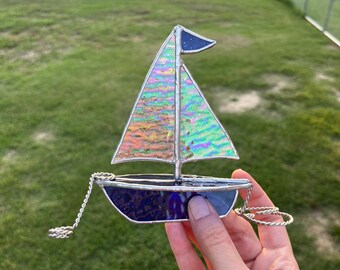 May include: A stained glass sailboat with iridescent blue and purple glass. The sailboat has a small blue sail and a silver chain attached to the bow.