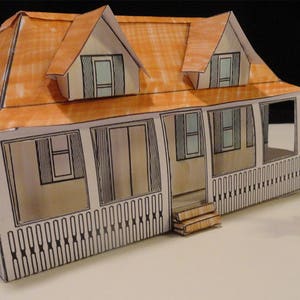 Könnte beinhalten: Ein Papiermodell eines weißen Hauses mit braunem Dach und einer Veranda. Das Haus hat mehrere Fenster und eine Tür. Vor dem Haus steht ein kleiner Schaukelstuhl.