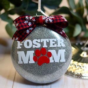 May include: Silver glitter Christmas ornament with a red and black plaid bow and the text "FOSTER MOM" in white with a red paw print.