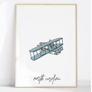 May include: A framed print with a minimalist line drawing of an early biplane in a light blue hue. The words "north carolina" are written in a cursive font below the aircraft. The artwork is set against a white background.