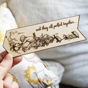 May include: A wooden bookmark with a laser-etched illustration of a group of animals pulling together. The text "and they all pulled together..." is above the illustration. The bookmark is held in a hand.