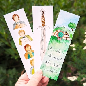 May include: Three watercolor bookmarks featuring characters and scenes from The Hobbit. The first bookmark shows four different illustrations of Bilbo Baggins. The second bookmark shows a watercolor illustration of a sword. The third bookmark shows a watercolor illustration of a hobbit hole with the text "In a hole in the ground there lived a hobbit."