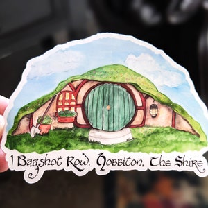 May include: A watercolor illustration of a hobbit hole with a green door and a red brick frame. The house is surrounded by green grass and a hill with a brown path leading to the door. The text "1 Bagshot Row, Hobbiton, The Shire" is written below the house.