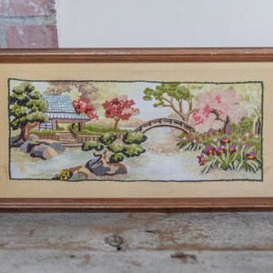 May include: A framed needlepoint embroidery depicting a Japanese garden scene with a bridge, trees, and a small house. The embroidery is done in shades of green, pink, and brown.