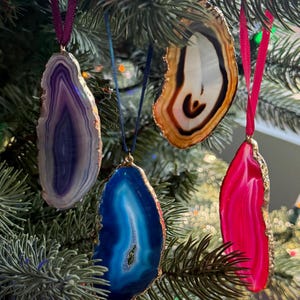 May include: A Christmas tree adorned with colorful agate slice ornaments. The ornaments are in shades of purple, blue, orange, and pink, each with a unique pattern and gold-colored edges. They hang from the tree with ribbons.