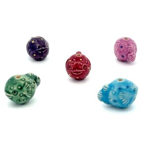 May include: Five ceramic pufferfish beads in various colors: purple, red, pink, green, and blue. Each bead features a detailed fish design with a smiling face and small holes for stringing. These beads are ideal for jewelry making.