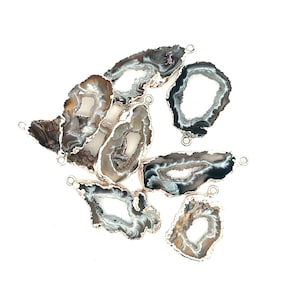 May include: A collection of agate slices with silver-colored metal edges and small loops for jewelry making. The slices feature a variety of colors, including brown, gray, white, and black, with a polished, smooth surface. The agate slices are arranged on a white background.