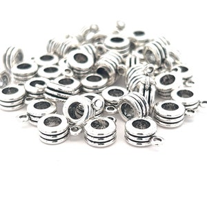 May include: A collection of silver-colored metal beads with a cylindrical shape. Each bead has a central hole and is adorned with horizontal grooves. Small loops are attached to the sides of the beads. The beads are scattered on a white surface.