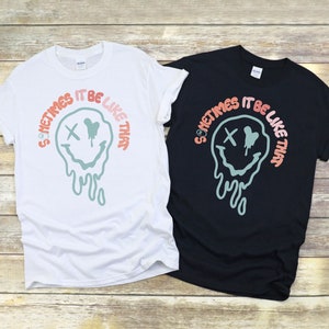 May include: Two t-shirts with a graphic design of a melting smiley face with an X for eyes and the text "Sometimes it be like that". The t-shirt on the left is white and the t-shirt on the right is black.