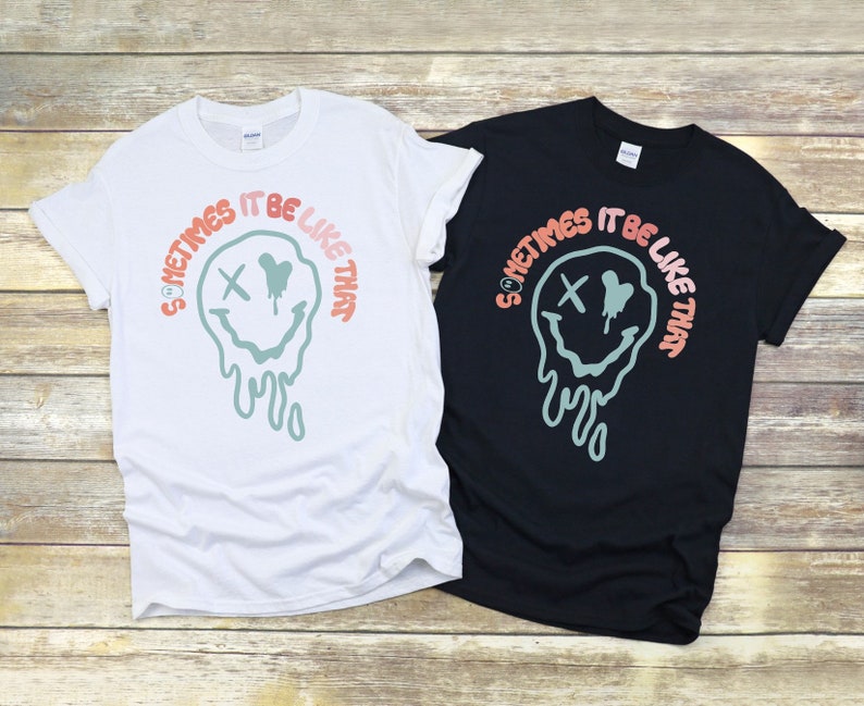May include: Two t-shirts with a graphic design of a melting smiley face with an X for eyes and the text "Sometimes it be like that". The t-shirt on the left is white and the t-shirt on the right is black.