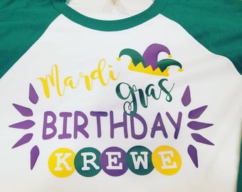 May include: White, green, and purple Mardi Gras birthday shirt with the text "Mardi Gras Birthday Krewe" and a jester's hat graphic.