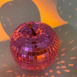 Puede incluir: Una bola de discoteca rosa con forma de manzana. La bola está hecha de pequeñas baldosas de espejo.