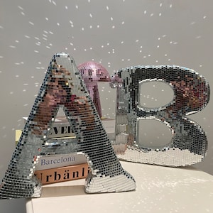 Peut inclure: Deux grandes lettres argentées, A et B, faites de carreaux miroir. Les lettres sont posées sur une surface blanche avec des livres empilés sous la lettre A. Les livres ont les mots "Barcelona" et "Urban" visibles.