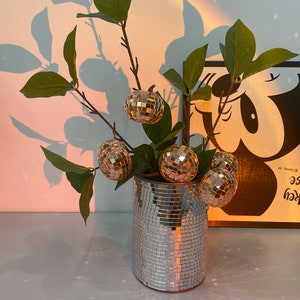 May include: A silver mirrored vase with a leafy green plant and five gold disco ball ornaments. The vase is sitting on a white surface. The background is a light blue wall with a shadow of the plant and a black and white illustration of Mickey Mouse with the text "Mickey Mouse" and "Disney" on the right side of the image.