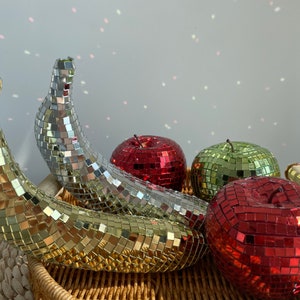 Può includere: Tre sculture di frutta decorative realizzate con piastrelle a specchio. Le sculture sono una mela rossa, una mela verde e due banane gialle. Le banane sono disposte in un cesto.