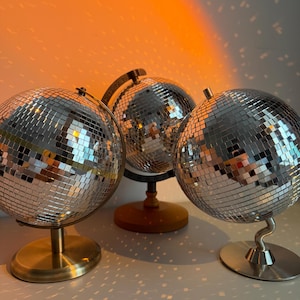 Puede incluir: Tres globos de discoteca con azulejos espejados, cada uno montado sobre un soporte diferente. Los soportes son de latón, madera y plata. El fondo es un degradado de naranja y blanco, con reflejos de luz.