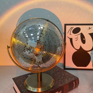 Puede incluir: Una lámpara de bola de disco dorada con una superficie de espejo. La lámpara está sobre un libro con una cubierta marrón. La lámpara proyecta una luz arcoíris en la pared detrás de ella.