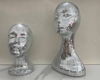 May include: Two silver mannequin heads covered in mirrored tiles. The heads are facing different directions. The head on the left is smaller than the head on the right.