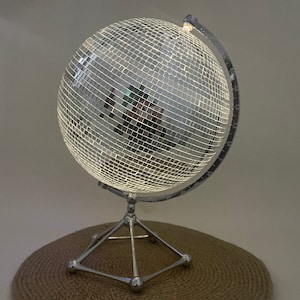 Puede incluir: Una lámpara de discoteca de globo plateado con una superficie de espejo y un soporte de metal. La lámpara está sobre una superficie tejida marrón.