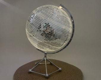 May include: A silver disco ball globe lamp with a mirrored surface and a metal stand. The lamp is on a brown woven surface.