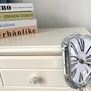 Puede incluir: Un reloj de espejo plateado con forma de reloj derretido. El reloj está sobre una cómoda blanca con una pila de libros encima. Los libros tienen los títulos "Evero is the Lubricant of Success", "Giorgio Morandi", "Barcelona" y "Urbänlike".