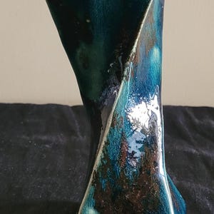 May include: A tall ceramic vase with a flared top and a twisted, angular body. The surface features a glossy, crackled glaze in deep shades of teal, navy blue, and dark brown, creating a complex, multi-tonal appearance with reflective highlights.