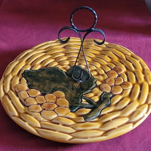 May include: A yellow ceramic serving platter with a woven basket design. The platter features a grape cluster and leaf design with a black metal handle. The colors are yellow, green, and brown.
