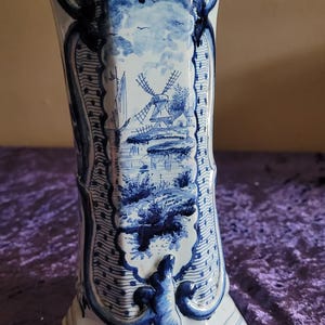 Può includere: Un vaso alto in ceramica blu e bianca con una scena di mulino a vento. Il vaso ha una parte superiore svasata e una base rettangolare. Il design include un mulino a vento, acqua e alberi. Il vaso è decorato con accenti floreali blu.