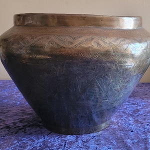 Könnte beinhalten: Eine große, antike Vase aus bronzefarbenem Metall mit aufwendigen Ätzmustern. Die Vase hat einen breiten, abgerundeten Körper, der sich zu einer kleineren Basis verjüngt. Der obere Teil weist ein dekoratives Band aus geätzten Mustern auf.