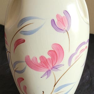 May include: A cream-colored ceramic vase with a crackle glaze finish, featuring two integrated side handles and a hand-painted floral motif in shades of pink, lavender, and light blue, with thin brown stems and leaves curving around the body.