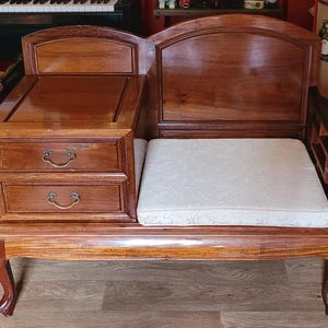May include: A wooden gossip bench with a polished brown finish, featuring a built-in side table with two drawers equipped with brass-coloured pulls. The seat is topped with a cream-coloured cushion featuring a subtle, light-coloured floral pattern.