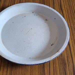 Peut inclure: Un plat à tarte en émail blanc avec un bord bleu. Le plat présente quelques petites taches de rouille à la surface.