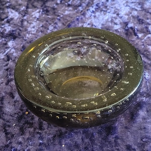 May include: A dark grey glass bowl with a rounded shape. The bowl has a wide rim and a shallow interior. Small air bubbles are visible throughout the glass, adding texture. The bowl sits on a purple surface.