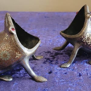 Puede incluir: Dos recipientes antiguos de metal con forma de rana. Los recipientes plateados tienen bocas abiertas, intrincados diseños florales y una gema roja en la parte superior. Se exhiben sobre una superficie púrpura.